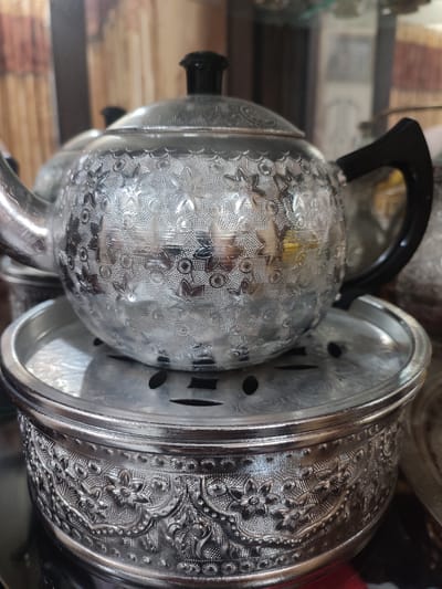 This media file features a close-up, eye-level shot of a decorative, metallic teapot and its stand. The teapot is silver-toned with intricate floral patterns etched all over its surface. It has a black lid and a black handle. The teapot rests on a circular, perforated metal stand, which in turn sits atop a wider, ornate silver-toned bowl that serves as the base of the stand. The bowl is also heavily decorated with embossed floral and scrollwork designs.
The setting appears to be indoors, possibly in a display cabinet or on a shelf, given the blurred background that shows hints of wooden textures and possibly other decorative items. The lighting is somewhat diffused, highlighting the metallic sheen and the detailed textures of the teapot and stand. There is no activity or event taking place; the objects are stationary. The image is focused on the craftsmanship and decorative elements of the tea set. No text is visible in the image. The overall impression is one of ornate, traditional craftsmanship.