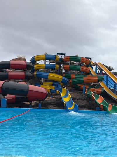 The image, rotated vertically, captures a dynamic water park scene under an overcast sky. No people are visible in the frame.

The left and central portions of the image are dominated by a complex network of colorful water slides. These slides feature distinct color combinations: intertwined segments of orange and green, alternating sections of bright yellow and blue, and darker red and black tubes. They twist and turn, supported by blue metal frameworks, and are integrated into what appears to be a multi-tiered, dark, rocky structure that mimics natural stone. Peeking from behind and between the slides are hints of green foliage, including palm trees, suggesting a tropical or warm-climate setting.

To the right, a large, light blue swimming pool occupies approximately one-third of the image. Its surface is textured with ripples and small waves, indicating movement. Several of the water slides terminate directly into this pool, with visible splashes of water where they meet the surface, suggesting active water flow. A segment of a yellow and blue slide is clearly seen entering the water. In the lower right corner, a red rope floats on the pool's surface near its edge.

The cloudy sky indicates a daytime setting, though not necessarily bright sunshine. The scene depicts the infrastructure of a vibrant water park, ready for recreational activity, consistent with a resort or leisure area like La Oliva, Spain. No visible text is present.
