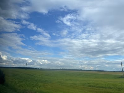 The image depicts a wide, green field under a dynamic sky with scattered clouds. A solitary wooden utility pole stands in the middle ground of the field. In the distance, a line of trees and some low-lying structures are visible, suggesting a rural or semi-rural area. The overall impression is one of an open, natural landscape. The location in the Капыльскі сельскі Савет, Belarus, confirms this rural setting. There are no people or animals visible in the image, and no actions or interactions are taking place. The environment is characterized by the vastness of the field and the dramatic sky.