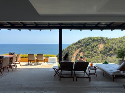 The image presents a serene outdoor-indoor living space overlooking the ocean and a lush green hillside in Puerto Marqués, Mexico. The foreground features a spacious concrete patio, with the view framed by an overhead roof structure and dark support pillars, indicating a sheltered veranda or terrace.
To the left, a large wooden dining table surrounded by woven chairs is set near a low, yellow-orange wall adorned with green foliage. Two additional beige chairs are positioned directly against this wall, facing the sea.
In the center and right, two individuals are relaxing on wooden lounge chairs by an infinity pool. Both are wearing distinctive green hats. One person, seen from behind, occupies a lounge chair in the mid-ground, facing the pool. The second person, largely obscured, is near the pool's edge, seemingly conversing with another individual whose green hat and arm are visible further right. A small wooden side table between the lounge chairs holds a green glass and some other items. Another lounge chair with neatly folded white towels rests closer to the pool.
Beyond the pool, a steep, verdant hillside covered in trees descends towards the expansive, calm blue ocean. The sky above is clear and bright, indicating a sunny day, likely midday or early afternoon given the distinct shadows cast on the patio. The architectural elements include the dark support beams and the detailed underside of the roof, with visible slats. The scene evokes a sense of relaxation and leisure amidst a beautiful coastal landscape. No discernible text is visible.