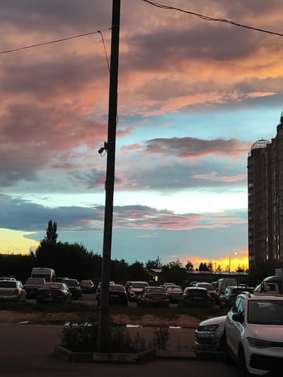 The image depicts a parking lot scene in the city of Shchyolkovo, Russia, during a sunset. The sky is filled with dramatic clouds, colored in hues of pink, orange, and blue. A tall, dark utility pole with wires runs vertically through the center of the frame, partially obscuring the view. In the foreground, several cars are parked. A white SUV is prominently visible on the right, facing left. Other vehicles, including sedans and what appears to be a van, are parked behind it and to the left, some with their headlights on. Beyond the parking lot, there is a dense line of trees silhouetted against the brightly lit sky. To the far right, a tall, multi-story brick building with arched windows and a domed structure on top dominates the skyline. Streetlights are visible in the distance, suggesting a residential or commercial area. No people are visible in the image. The overall impression is of an urban environment at dusk.