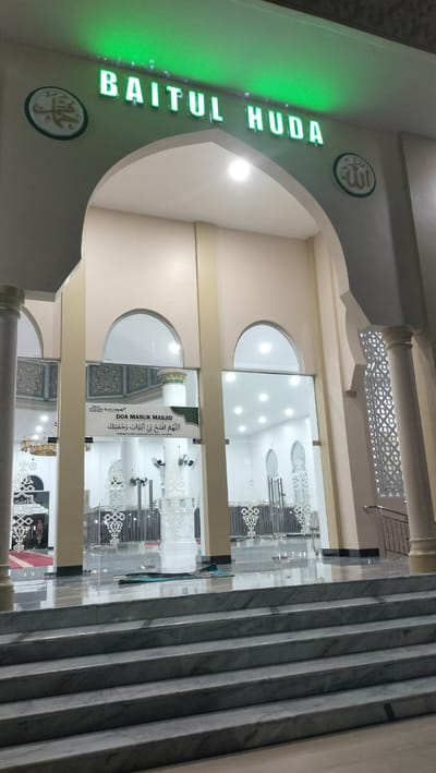 The image displays the brightly lit entrance of a mosque named Baitul Huda, located in Mee Tanjong Usi, Indonesia, captured during the evening or night. A large archway above the entrance features the mosque's name "BAITUL HUDA" in prominent green glowing letters. On either side of the name, circular emblems showcase Islamic calligraphy: the name of Muhammad (محمد) on the left and the name of Allah (الله) on the right.
A set of polished, light-colored marble steps leads up to the mosque's main entrance, which consists of tall glass doors. Through these doors, the well-illuminated interior of the mosque is visible, featuring reflective floors, decorative white screens, and bright ceiling lights. A green prayer mat is visible on the floor just inside the entrance.
A banner hanging inside, visible through the glass, displays the text "DOA MASUK MASJID" (Prayer for entering the mosque). Below this, the Arabic prayer "اللَّهُمَّ افْتَحْ لِي أَبْوَابَ رَحْمَتِكَ" (Oh Allah, open for me the gates of Your mercy) is printed, along with its Indonesian translation. A small "BA Bank Aceh Syariah" logo is also present on the banner. The architectural details include substantial pillars and ornate, geometrically patterned latticework on the right side. No individuals are present in the scene.