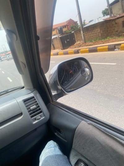 The image shows a view from inside a vehicle moving along a road in AlakIa, Nigeria. The vehicle's side mirror reflects the street, including a building and a curb with alternating yellow and brown painted blocks. The interior of the vehicle is visible, including the air vent, part of the door, and a denim-clad leg.
