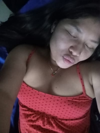A woman with long, dark hair reclines with her eyes closed, lips pursed in a kiss. She wears a coral-colored top with small, dark polka dots and delicate spaghetti straps. A gold necklace with a small, decorative pendant rests on her chest. The background is dark, possibly a blue or dark-colored blanket. The lighting is somewhat dim, casting shadows and emphasizing her facial features.