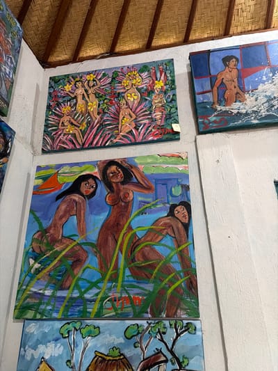 The image shows several paintings hanging on a white wall in what appears to be an art gallery or shop. The ceiling above is made of woven natural material. The paintings depict human figures and natural scenes.
One painting at the top left shows several nude figures of women with yellow flowers in their hair and on their bodies, surrounded by lush pink and green foliage.
To the right of this, a painting shows a muscular, nude male figure emerging from turbulent white water, with a dark blue background and red accents.
Below these, a large painting occupies the center of the frame and shows three nude female figures in water with tall green grass. One figure on the left looks directly at the viewer, while the figure in the center has her hand on her head and the figure on the right looks away. A small orange boat is visible in the background.
Below this, another painting depicts a thatched-roof structure with trees and green foliage against a blue sky with white clouds.
The overall impression is of an art display featuring figurative and landscape paintings with a vibrant, somewhat naive style. The location is identified as Tejakula, Indonesia.