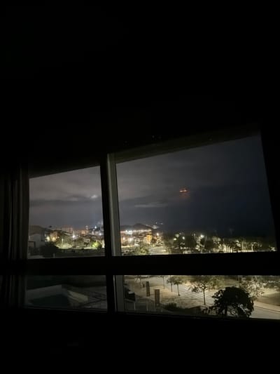 The image displays a nighttime view of Villajoyosa/la Vila Joiosa, Spain, observed from an elevated interior perspective through a large, multi-paned window. The immediate foreground is dark, revealing a light-colored curtain on the far left and the dark frame of the window. Through the window, the cityscape is visible on the left, illuminated by numerous artificial lights from buildings, streets, and residential areas, suggesting a dense urban environment extending towards hills in the distance. A partial view of a swimming pool is discernible below the left section of the window. On the right, a paved area or road with trees and streetlights is visible, leading towards the coastline. Beyond the land, the dark expanse of the Mediterranean Sea is apparent under a cloudy night sky. A distinct cluster of four to five bright orange lights is visible on the dark horizon over the sea, likely originating from a distant vessel or offshore structure. No individuals or specific actions are discernible in the scene.