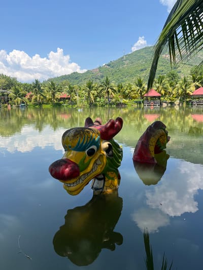 This is a photograph of a decorative dragon head and body emerging from the water of a tranquil lake. The dragon is brightly colored with yellow, red, green, and blue. Its head is in the foreground, with the body visible further back in the lake. The lake is surrounded by lush palm trees and verdant hills, with several red-roofed structures visible along the shoreline. The sky is a clear blue with scattered white clouds, which are reflected in the water. The scene appears to be a scenic resort or park area in Suối Tân, Vietnam.