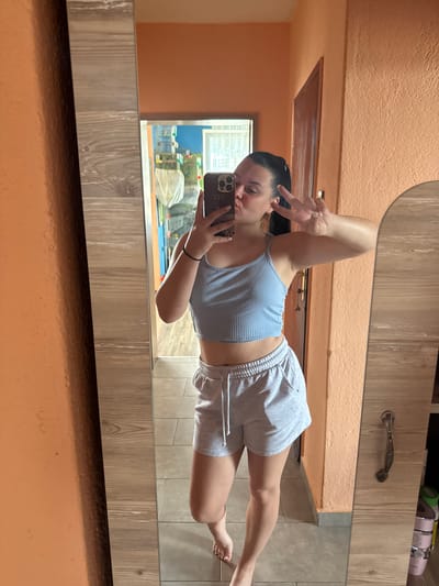 A young woman with dark hair in a ponytail is taking a mirror selfie in what appears to be a residential interior. She is wearing a light blue ribbed tank top and grey shorts. She is puckering her lips and holding up two fingers in a peace sign. She is holding a smartphone in her right hand, which is angled to capture her reflection. The background includes a wooden-framed mirror, an orange-painted wall, a door with an ornate handle, and a tiled floor. In the reflection of the mirror, there are some shelves with items on them, and a window. The overall impression is that of a casual, domestic setting. There is no specific indication of the location being in the city of Rogosh, Bulgaria.
