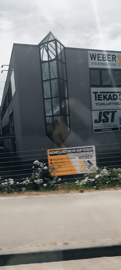 This is a photograph of a modern commercial building located in Winterbach, Germany. The building has a grey facade and features a prominent glass-enclosed structure on its upper portion. Several signs are visible on the building's exterior. One sign reads "WEBER STEUERBERATUNG," indicating a tax consulting firm. Another sign displays "TEKADÖ TURLUFTSCH," likely related to air ventilation or similar services. A third sign shows "JST" with the text "J.S.T. DEUTSCHLAND GMBH VERSAND," suggesting a shipping or distribution company.
In front of the building, there is a black fence with a row of white flowering bushes. A yellow and white sign is attached to the fence, featuring the text "KEINE LUST MEHR AUF STAU?" which translates to "No more desire for traffic jams?". Below this, further text and logos are present, including "WEBER STEUERBERATUNG". The ground in front of the fence appears to be a paved area, possibly a road or sidewalk. The sky is overcast with some visible light rays. The overall impression is of a commercial district under a cloudy sky.