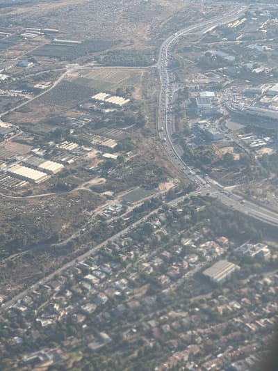 This aerial photograph captures a sprawling urban and semi-urban landscape. The scene is dominated by roads, buildings of various sizes and architectural styles, and patches of vegetation. The perspective is from a high vantage point, likely an aircraft, looking down at a tilted angle.

The subjects are primarily man-made structures and infrastructure. We see a highway with multiple lanes of traffic, indicating significant transportation activity. Alongside the highway, there are residential areas with densely packed houses, many featuring red-tiled roofs, suggesting a developed neighborhood. Further away, there are larger industrial or institutional buildings with flat roofs, some appearing to be warehouses or factories. The presence of agricultural fields, characterized by neat rows of crops or greenhouses, suggests a mix of urban and rural land use. Numerous cars are visible on the roads and in parking areas, implying a daytime scene.

The weather appears to be clear, with good visibility, allowing for a detailed view of the terrain and structures. The sunlight casts shadows, though they are not particularly pronounced, suggesting it is not early morning or late afternoon. The overall impression is one of a functional and populated area, with daily life in progress. No specific event is clearly discernible, but the general activity suggests a normal day.

Location cues, such as the style of buildings and the layout of the urban areas, combined with the provided location context of Modi'in Illit, suggest this is likely a settlement in the Palestinian Territories. There is no visible text in the image.
