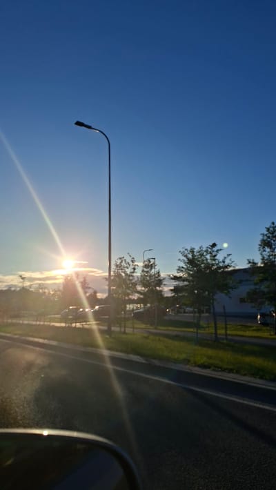 The image captures a road scene at dusk, likely in the vicinity of Harkujärve, Estonia. The sky is a clear, deep blue, transitioning to lighter shades near the horizon. The sun is low in the sky, casting a bright glare and prominent sunbeams across the left side of the frame, partially obscured by thin clouds. A tall, dark streetlamp stands silhouetted against the sky in the left half of the image. To the right, a row of trees with green foliage line the roadside. Beyond the trees, a building with a light-colored facade and darker windows is visible. A fence runs along the edge of the property. The road surface, asphalt, is dark and shows tire marks and a white lane marking. The foreground includes the curved edge of a car's rearview mirror, suggesting the photo was taken from inside a vehicle. Several cars are parked or driving on the road in the mid-ground, and the overall impression is one of a suburban or commercial area on the outskirts of a town.