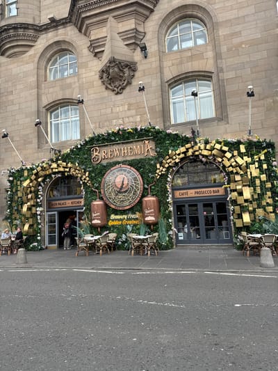 The image shows the exterior of a building, identified as "BREWHAMIA", located in the city of Edinburgh, United Kingdom. The building's facade is made of stone and features arched windows. The entrance is heavily decorated with artificial greenery, flowers, and golden geometric shapes. A large clock with a brewing-themed design is prominently displayed. Outdoor seating with tables and chairs is arranged in front of the entrance, with some patrons seated at the tables. The street in front of the establishment is paved, with white lines indicating traffic lanes. Several people are visible, including patrons seated at tables and individuals entering or exiting the building. The overall scene depicts a bustling outdoor dining and drinking establishment.