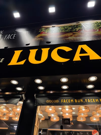 The image displays the illuminated facade of a LUCA establishment, located in Agigea, Romania.

The upper section of the facade features a large billboard or banner with a high-resolution image of a sandwich, depicting a baguette-style bread filled with lettuce, cheese, and a fried protein, likely chicken. Partial white text, including "N." and "FACE", is visible to the left of the sandwich image. Above this banner, two square, bright white light fixtures are mounted on a dark ceiling.

Below the billboard, a prominent black sign displays the name "LUCA" in large, illuminated yellow capital letters. Beneath the "LUCA" sign, a dark horizontal panel is fitted with multiple evenly spaced, circular recessed spotlights.

The lower part of the facade consists of two distinct storefront sections. Each section features a black roll-down awning with yellow text. The left awning shows partial text ending in "...traditie.ro", and below it, "RĂCORITOARE" (refreshments) is visible. The right awning displays the phrase "FACEM BUN, FACEM F..." (We make good, we make fast/fresh), a common slogan for the chain. A white sign hanging below the right awning states "PROGRAN NON STOP" (non-stop schedule). Visible through the storefront windows are numerous interior pendant light fixtures, each with a distinctive woven, basket-style shade.