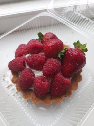 A close-up, top-down view shows a single fruit tart presented in a clear plastic container. The tart has a golden-brown, fluted crust filled with white cream. It is generously topped with fresh raspberries and strawberries. The berries are vibrant red, with some strawberries showing their green leafy tops. The tart appears to be part of a pre-packaged dessert, possibly purchased from a bakery or store in Domodedovo, Russia. The lighting suggests it is daytime, and the overall mood is one of simple indulgence. There is no visible text in the image.
