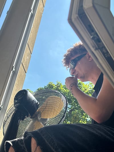 The image shows a person, likely male, with curly hair and wearing sunglasses, looking down at a fan. He is wearing a black t-shirt and ripped black jeans. The scene appears to be outdoors, with a clear blue sky and green trees visible in the background. The sun is shining brightly, suggesting it is daytime. The person is holding his hand near his mouth, and a bracelet is visible on his wrist. The fan is black and has a mesh grill. The perspective of the photo is from below, looking upwards. A light-colored ledge or structure is visible in the foreground. The overall impression is a candid shot of someone enjoying a warm day.