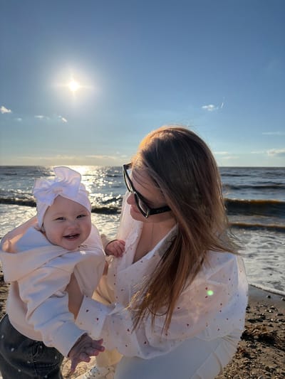 In Port-Katon, Russia, a woman wearing sunglasses and a white shirt with heart patterns holds a baby on a beach. The baby, wearing a white bow and a cream-colored hoodie, smiles at the camera. The sun shines brightly in the blue sky, reflecting on the water, and waves lap against the shore. The beach is covered in sand and small stones.