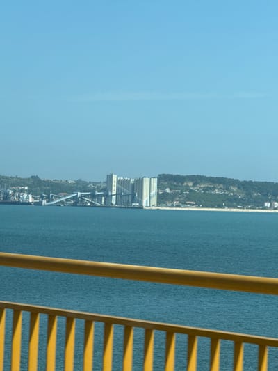 The image captures a wide view of the Tagus River estuary on a clear, sunny day. In the distance, a prominent industrial complex is visible along the coastline, characterized by large, cylindrical silos and a network of elevated conveyor belts. These structures are set against a backdrop of a verdant, hilly landscape dotted with buildings, suggesting a coastal town or city. The water in the foreground is a deep blue, with a gentle ripple. In the immediate foreground, a bright yellow railing, likely part of a bridge or elevated walkway, bisects the lower portion of the frame. The railing, composed of a thick horizontal bar and vertical slats, casts shadows on the water. The overall impression is one of a working port or industrial area adjacent to a populated coastal region. Based on the visible industrial structures and coastal geography, this scene is consistent with the industrial port area of Barreiro on the south bank of the Tagus River, facing Lisbon and Oeiras.