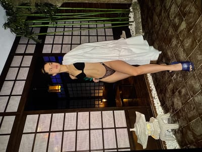 A woman in a black bikini and a leopard print bottom is posing on a tiled floor in front of a Japanese-style screen with bamboo stalks and paper windows. She is wearing blue flip-flops. The scene appears to be outdoors at night, with a lantern visible in the background. There is a stone decorative object on the right side of the image. The location of this image is not in Denver, United States.