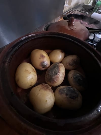 The image depicts a close-up view within a kitchen environment in Kfar Vitkin, Israel, focused on a dark brown, possibly earthenware, pot containing eight small, whole potatoes. Several potatoes exhibit light yellow skins with minor blemishes, while others show areas of significant charring or dark discoloration on their surfaces. In the background, an out-of-focus, lidded earthenware pot, possibly a tagine, sits behind the potato pot. To the right, a black frying pan rests on a gas stove burner, actively cooking several pieces of meat, from which steam or smoke is visibly rising. The metal grate of the gas stove is discernable beneath the pan. A roll of white paper towels is located on a countertop adjacent to the stove. A tiled wall and a dark, reflective surface, likely an appliance or cabinet, are visible in the ambient background. No individuals are present in the scene.