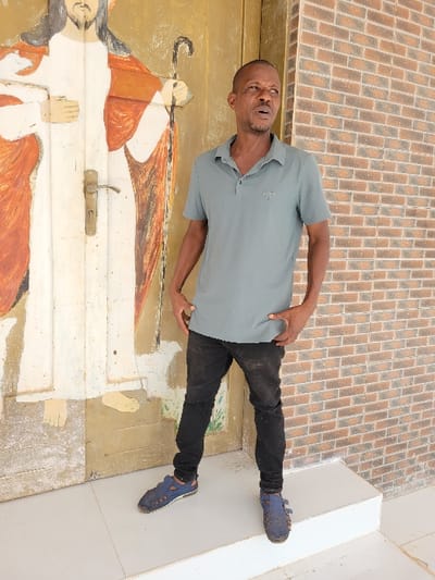 In Ebenebe, Nigeria, an adult male stands on a light-colored, possibly tiled or concrete, elevated platform. He wears a teal-grey short-sleeved polo shirt, faded black denim jeans, and dark blue closed-toe sandals with visible stitching. His hands are positioned near his hips, and his gaze is directed towards the viewer's right. The man has short, dark hair, light facial hair, and a slightly open mouth. To the left of the frame, behind the man, is a light brown wooden door featuring a worn full-length painting of a robed figure, presumably Jesus Christ, holding a shepherd's crook. A brass-colored door handle is present on the painted door. To the right of the frame, behind the man, a section of a reddish-brown brick wall with white mortar is visible.
