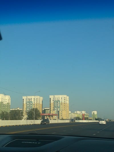 The image depicts a street scene in a city, likely residential, with modern apartment buildings visible in the background. The buildings are tall and multi-story, with varied architectural styles and colors, including white, yellow, and green. One of the buildings has a red and yellow sign that reads "SINOOIL," indicating a gas station or related business. The road in the foreground is paved and has yellow and white lane markings. Several cars are on the road, including a dark-colored van and a white sedan. A white fence with yellow accents runs along the side of the road. Trees and greenery are present between the road and the buildings. The sky is clear and blue, suggesting daytime. The view appears to be from inside a vehicle, as the bottom of the image shows part of a car's dashboard. The scene is characteristic of urban development in Kazakhstan, with the presence of the "SINOOIL" brand and the architectural style of the buildings being consistent with areas in cities like Nur-Sultan (Astana).