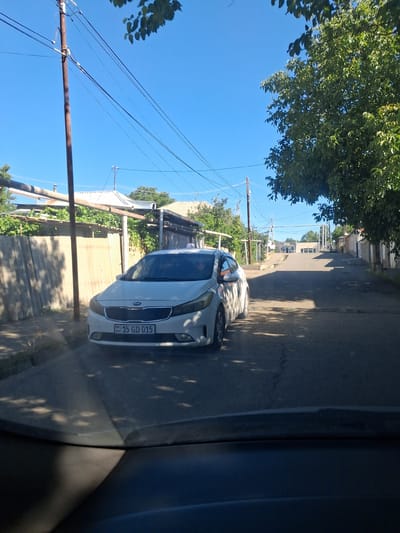 The image shows a white Kia sedan parked on a street in Yeghegnadzor, Armenia. The car has a license plate that reads "15 GD 015". The street is paved and appears to be a residential area, with houses and trees visible in the background. The sky is clear and blue. The image is taken from the perspective of a driver looking out of the windshield of another vehicle.