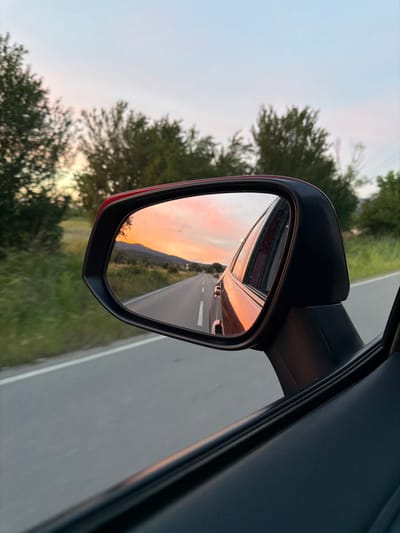 This is a medium-angle, close-up shot of a car's side-view mirror, taken from inside the vehicle. The mirror reflects a road stretching into the distance, bordered by green vegetation. The sky above the road is a soft, pale blue, transitioning into vibrant hues of pink and orange as it meets a mountain range on the horizon, indicative of a sunset. The reflection also captures the rear of the car, a sliver of its red exterior visible along the left edge of the mirror's frame.
Outside the mirror, the car's interior is visible on the right, a dark, textured surface. To the left of the mirror, out of focus, are trees with green foliage against the same pale sky. The overall scene evokes a sense of travel and the peaceful beauty of a sunset viewed from the road. The time of day is clearly sunset. The weather appears clear, as indicated by the visibility of the sky and landscape. The image has a slightly reddish tint due to the sunset. No visible text is present in the image.