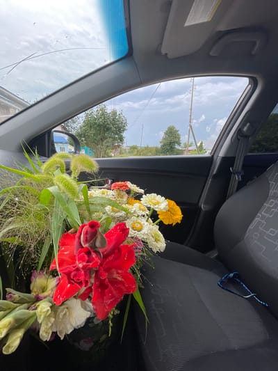 A bouquet of flowers, including red gladioli, white and yellow zinnias, and green foliage, is placed on the passenger seat of a car, seen from the interior. Outside the car window, a cloudy sky with visible cracks in the windshield is visible, along with trees, houses, and utility poles in the village of Perevoznoye, Russia. A pair of blue and white beaded glasses rests on the car seat next to the flowers.