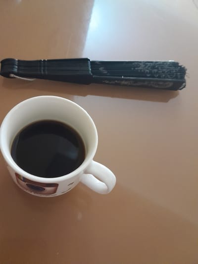 A black fan and a white mug of coffee sit on a brown, reflective surface. The fan is closed and standing upright, to the left of the mug. It has decorative etchings on its surface and a small white string attached to its base. The mug is positioned to the right, angled slightly upwards and to the left. It is white with a handle and contains dark liquid, presumably coffee. There are some faint, colorful patterns on the side of the mug. The background is a plain, smooth surface with subtle variations in color and some light reflections. The lighting suggests it could be daytime, with light coming from the upper left. There is no indication of weather or specific emotional cues. No legible text is visible on the fan or mug. The setting appears to be indoors, possibly on a table.
