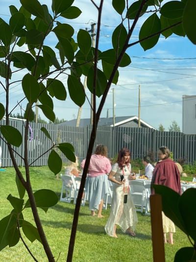 The image depicts an outdoor gathering at a private residence in Simagino, Russia. In the foreground, the branches and leaves of a tree obscure the view. In the midground, several individuals are present, some seated at a white table set for dining. A woman in a cream-colored dress and another in a maroon jacket are standing. The background shows a grey vertical slat fence, some trees, and residential buildings. The overall scene suggests a social event taking place in a backyard on a clear day.