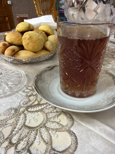 A glass of amber-colored liquid, likely tea, sits on a saucer with a decorative silver rim, placed on a white tablecloth with intricate lace trim. Beside the glass, a crystal dish holds an assortment of golden-brown baked goods, possibly cookies or small breads. In the background, a crystal decanter filled with ice cubes is visible, along with a blurred bottle and a hint of wooden furniture. The scene appears to be a domestic setting, possibly a dining table in Balakənd, Azerbaijan, set for a tea time gathering.