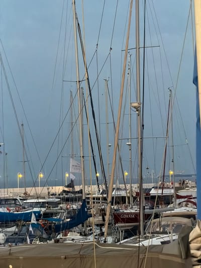 The image depicts a marina in Sopot, Poland, likely during twilight or overcast conditions, as indicated by the muted lighting and the color of the sky. Numerous sailboats with their masts and rigging dominate the foreground and midground. The boats are docked closely together, creating a dense arrangement. Several yellow-orange streetlights are visible, illuminating parts of the marina.
One prominent sailboat in the mid-right of the image is dark red with white lettering that reads "GDANSK." Another sailboat further back has a white flag with red lettering that appears to say "REMEDIC." There are also other boats of various sizes and colors, including white and blue hulls and sails.
In the background, a walkway or pier is visible with a few people walking. The sea or body of water is beyond the marina, with distant lights suggesting the presence of a shoreline or another part of the city. The overall impression is one of a busy, albeit somewhat subdued, harbor.