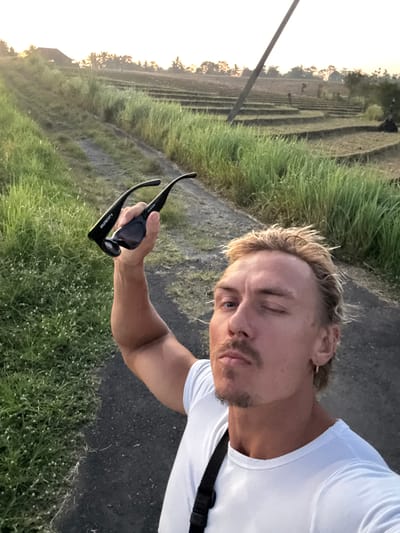 A selfie of a male with blonde hair and a short beard, wearing a white t-shirt. He is holding black sunglasses in his right hand. The background features green grass and what appears to be rice terraces, indicating a location in Selemadeg Timur, Indonesia. A pole stands to the left, and the light suggests it is either early morning or late afternoon.