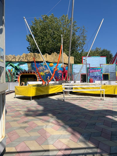 This media file depicts an outdoor amusement park scene in Golubitskaya, Russia, under a clear blue sky, suggesting a sunny day. The main subjects are the attractions themselves, including a "Mirror Maze" (ЗЕРКАЛЬНЫЙ ЛАБИРИНТ) and what appears to be a "Parrot World" (МИР ПОПУГАЕВ). The Mirror Maze attraction features a metallic, reflective facade with colorful signage. Nearby, a bungee trampoline setup is visible with metal poles and elastic cords. The ground is paved with a pattern of red, grey, and beige interlocking bricks, creating a visually interesting walkway.
The scene is set up for entertainment, with bright colors and playful imagery on the attraction facades. The "Parrot World" entrance is adorned with a vibrant mural depicting a jungle scene with exotic birds, fish, and marine life, including what looks like a Spider-Man figurine integrated into the design. Orange netting or rope hangs across the attraction. The overall atmosphere is that of a lively amusement area. The time of day is likely daytime, given the bright sunlight casting distinct shadows on the paved ground. The location is identifiable as Golubitskaya, Russia, indicated by the text on the signage. No people are clearly visible in the frame, focusing the attention on the attractions and the setting.