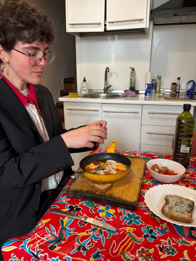 A person in a black blazer sits at a table covered with a red floral tablecloth. On the table is a frying pan filled with food, two slices of toast, a bowl of tomatoes, a knife, and a bottle of oil. The person is holding a fork near the pan. Behind the table is a kitchen with white cabinets and a sink.