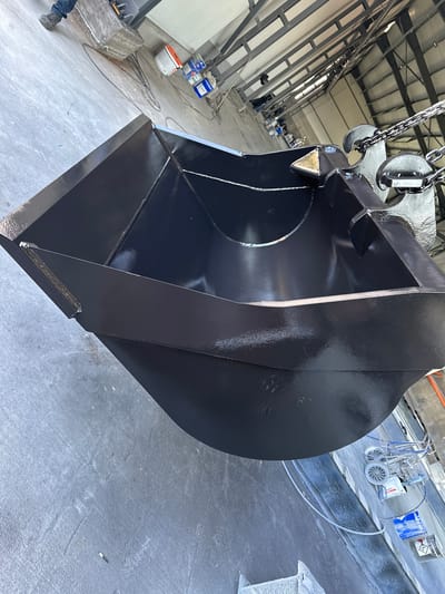 The image shows a large, black, recently painted excavator bucket suspended by chains in what appears to be an industrial setting. The location seems to be a factory or workshop interior, possibly in Vedrare, Bulgaria, based on contextual clues. The bucket dominates the foreground, showcasing a glossy, newly applied coat of black paint. The background reveals industrial building elements like support beams, concrete floors, paint sprayers, and various containers, potentially paint buckets. No people are clearly visible, but indistinct figures exist in the background, suggesting a workforce presence. The overall scene suggests a stage in the manufacturing or refurbishment process of the excavator bucket.