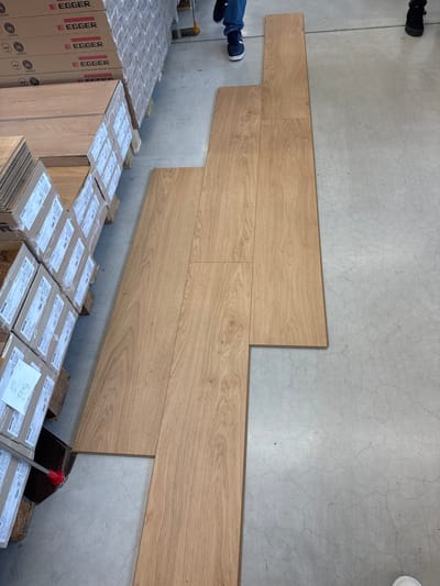 A medium-angle, eye-level indoor shot shows flooring installation in progress on a light gray concrete floor. Four rows of light brown wood-look planks are laid in a staggered pattern against the wall, occupying the upper right quadrant of the frame. The bottom left quadrant is filled with boxes of flooring. The “EGGER” logo is printed on vertical brown boxes, stacked on a wood pallet. Horizontal, beige boxes with paper labels, partially obscuring the writing, are also visible. A white piece of paper with hand-written text is visible on the top of one of the beige boxes near the lower right. The location of the image is unknown as well as the actions or interactions of people in the image.