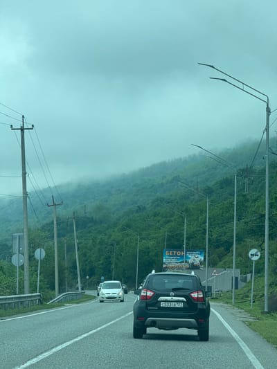 The image shows a paved road winding through a misty, mountainous area. A black Nissan Terrano SUV is driving on the right side of the road with the license plate "c533cb 123." A white car is visible in front of the SUV. On the side of the road, there are several utility poles with power lines, as well as road signs and a billboard advertising concrete ("БЕТОН" in Russian). The backdrop is a forest-covered mountain shrouded in fog. The location appears to be a road through a mountainous region in or near Sukhumi, Abkhazia. The cars and signage indicate active transit in the area.