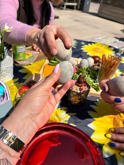 The image captures a festive outdoor gathering, likely an Easter celebration, set in Monistrol de Montserrat, Spain. The scene is viewed from above, focusing on hands and a table laden with food and decorations under bright sunshine.
Several hands are prominently featured, engaged in an egg-tapping game, a traditional Easter activity. In the foreground, two hands hold grey, speckled eggs, appearing to clink them together. One hand has light pink nail polish, while the other has natural nails. Another hand from the upper right, adorned with vibrant blue nail polish, also holds a similar grey egg. Further back, another pair of hands displays an alternating pattern of bright yellow and blue nail polish.
The table is covered with a dark blue cloth featuring large, cheerful yellow sunflower patterns, enhancing the spring and festive atmosphere. Various items are spread across the table: a glass filled with thin breadsticks, small round fruits or olives, fresh green lettuce, and slices of orange food, possibly cheese or fruit, arranged on wooden serving boards. A decorative egg-shaped bowl, intricately patterned, holds a dip or salad garnished with what looks like dried berries. A shiny red metallic plate is visible on the right side of the frame, along with a white lace-patterned cup or holder and a small white flower with a green stem, adding to the decorative elements.
A person wearing a light pink long-sleeved top is visible on the left. On the right, a wrist adorned with a silver and gold watch is seen. The strong sunlight casts distinct shadows, indicating a bright daytime setting. The overall mood conveyed is one of joy, tradition, and communal celebration in an inviting outdoor environment. No clearly legible text is visible on any objects in the image, though a barcode can be seen on a green can in the background.