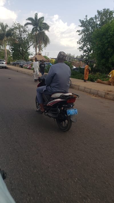 A person wearing a gray outfit and a patterned cap is riding a motorcycle on a paved road. The motorcycle has a blue license plate. On the left side of the road, several people are walking along the side of the road. A tall palm tree and other greenery are visible. Cars are parked behind the people. On the right side of the road, there are more trees and shrubs. The road appears to be in Hayin Banki, Nigeria.