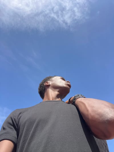 A low-angle, waist-up shot shows a person looking upwards against a bright blue sky with scattered white clouds. The person, viewed from the chest up, is wearing a black, ribbed t-shirt. Their right arm is bent, with their hand resting on their shoulder, revealing a black and silver braided bracelet on their wrist. The skin tone is dark. The image was taken in Sant Adrià de Besòs, Spain.