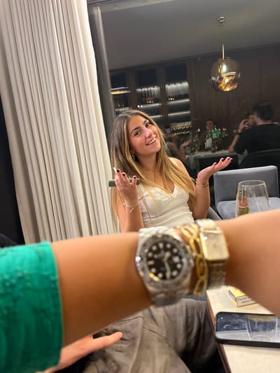 An indoor scene depicts a social gathering, likely within an upscale restaurant or bar. The specific location within Lordelo do Ouro e Massarelos, Portugal, cannot be determined from visual evidence.
In the foreground, a person's left arm, wearing a green sleeve, is positioned prominently, displaying a silver-toned wristwatch with a black dial and decorated bezel, a gold-toned chain-link bracelet, and a second gold-toned rectangular wristwatch. The arm is holding a smartphone, suggesting it belongs to the photographer. On a light-colored table to the right, a white and yellow cigarette package with the text "FUMAR" is visible, along with a dark smartphone and a clear glass.
The mid-ground features a young woman with long, light brown hair, smiling directly at the camera. She wears a white v-neck top and multiple thin bracelets on both wrists. Her left hand, with red painted fingernails, holds a dark rectangular object, possibly a vape pen or small device. Her right hand is open, palm up, in a gestural pose.
The background is dimly lit and out of focus, revealing several other patrons seated at tables. Decorative elements include tall, light-colored pleated curtains to the left, a gold-toned spherical pendant light fixture, and a dark wood shelving unit or wall paneling illuminated by strip lighting, possibly displaying bottles. Grey upholstered seating and tables with light surfaces are visible. The overall environment suggests a modern, relaxed, indoor social setting.