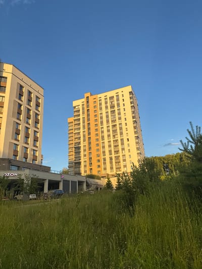 This media file depicts an outdoor scene with a clear blue sky. Dominating the frame are two modern residential buildings. The taller building on the left is a light-colored apartment complex with many windows and balconies, featuring an orange accent on its facade. To its right, a second, slightly shorter building is visible, also with numerous windows and a beige exterior. In the foreground, tall, lush green grass fills a significant portion of the image, suggesting a natural, unkempt area adjacent to the urban development. A few trees are also present, including what appears to be a pine tree in the upper left corner and denser foliage in the mid-ground.
The lighting suggests it is late afternoon or early evening, with a warm golden hue cast on the buildings. The weather appears to be clear and pleasant. On the lower building, the text "OZON" is visible in a stylized font, indicating a commercial presence. The overall impression is of a contemporary residential area in Koltsovo, Russia, where urban architecture meets nature. There are no people visible in the image, and the scene is static, conveying a sense of calm and everyday life.