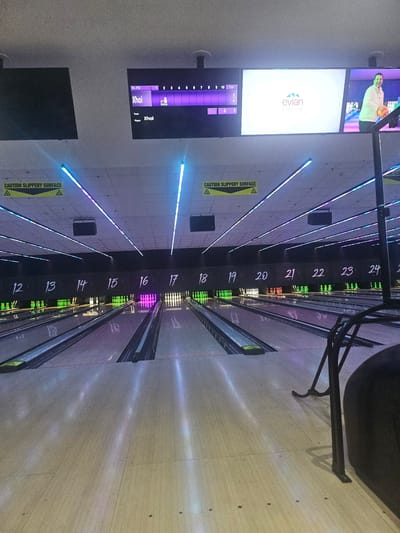 The image depicts an indoor bowling alley with multiple lanes. The lanes are numbered from 12 to 24, and some lanes have pins set up. There are screens displaying scores and player information, with the name "Xhoi" visible. One screen features the Evian logo, and another shows a person holding a bowling ball. The environment is dimly lit, with neon lights running along the ceiling. There are "CAUTION SLIPPERY SURFACE" signs visible. A bowling ball return mechanism and a black chair are in the foreground. The scene is devoid of any specific architectural details that would allow for precise location identification within Gloucester, United Kingdom.