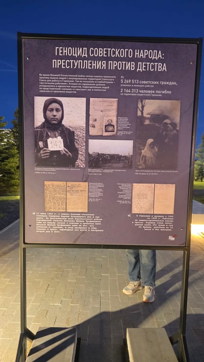Here's a detailed description of the image:
**Location:** Likely Zaitsevo (Verevskoe S/P), Russia. This is based on the Russian text in the image.
**Description:**
The image shows a rectangular information panel, likely part of a memorial or historical display, standing outdoors. The panel details the genocide of the Soviet people, specifically focusing on crimes against children during the Great Patriotic War (World War II).
**Content of the Panel:**
* **Headline:** The panel's title is "Genocide of the Soviet People: Crimes Against Childhood."
* **Text:** The text describes the practice of the German occupiers during World War II, who deported Soviet citizens from occupied territories to Germany as forced laborers ("Ostarbeiters"). It mentions the poor conditions, and the ideological motivations based on the Nazi's division of people into "lower" and "higher" races. Statistics are given regarding the number of Soviet citizens deported and killed.
* **Images:** The panel contains several black and white photographs and scanned documents as visual evidence to support the narrative. These include:
* A photograph of a child with a sign showing their assigned number "142".
* Various historical documents in Russian, including work permits, reports about murders committed by Nazi troops, and personal autobiographies.
* Images of a village being burned.
* **Citations:** There are citations referring to various archives (e.g., GANIRO, GAVO).
**Other Details:**
* In the bottom right corner, there is a portion of a person's lower legs and feet visible, wearing jeans and sneakers, implying someone is standing nearby and looking at the display.
* The panel is mounted on a metal frame.
* The surrounding environment seems to be a paved area, possibly a park or memorial site, with some greenery visible in the background.
**Overall:** The image appears to be part of an outdoor exhibit or memorial, likely focused on the historical impact of World War II on the Soviet people, with an emphasis on the crimes committed against children.