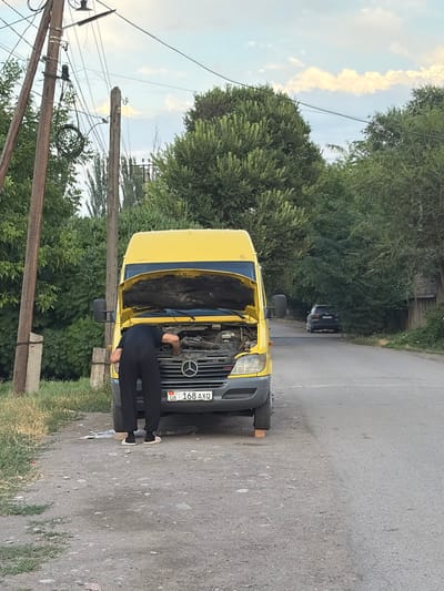 A person is working on the engine of a yellow Mercedes-Benz Sprinter van. The van's hood is open, and the person is bent over, appearing to be inspecting or repairing the engine. The license plate of the van is visible and reads "KG 168 AXQ". The scene takes place on a paved road in a somewhat rural or suburban area, with trees and vegetation lining the sides. In the background, there is a dark-colored SUV parked on the side of the road and a wooden building. Utility poles with wires are prominent in the left foreground, suggesting the presence of infrastructure. The lighting suggests it is daytime, possibly late afternoon, given the soft light and the presence of shadows. The ground is a mix of asphalt and gravel. The general environment suggests a location outside of a dense urban center, potentially on the outskirts of the city of Kant, Kyrgyzstan.