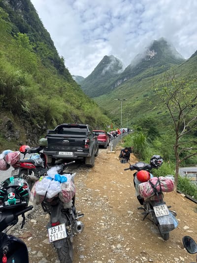 A line of parked vehicles, primarily motorcycles heavily laden with luggage, and a dark-colored pickup truck, are oriented along a dirt road. The vehicles are positioned roadside to the right, with a steep, verdant mountain rising to the left of the frame. Overcast skies and mist-shrouded mountain peaks are in the background. The license plates on the vehicles indicate that the location is likely Yên Minh Commune, Vietnam. People are seen standing and walking along the road between the vehicles.