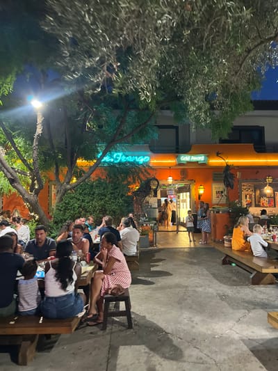 The image displays the illuminated outdoor dining area of Marisqueira e Restaurante Sr. Frango, located in Almancil, Portugal, during nighttime hours. The restaurant building exhibits an orange facade with a prominent green neon sign reading "Sr. Frango" and a "Gold Award" sign positioned above the main entrance. Approximately 35 individuals, comprising adults and children, are present. Most are seated at multiple dark-stained wooden picnic-style tables with integrated benches, which are arranged on a concrete ground surface. Table settings include plates, glassware, and beverage bottles. The outdoor space features several trees, some adorned with string lights around their trunks, and various planted shrubs. Illumination is provided by a tall street lamp on the left, the restaurant's exterior lighting, and the neon signage. Near the main entrance, a large, dark, bird-like sculpture is visible. Individuals at the tables are engaged in dining and conversation, while a small group of people are positioned near the brightly lit entrance, some appearing to enter or exit the establishment.