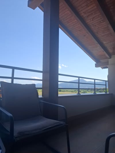 The image captures a view from a covered outdoor seating area, likely a balcony or patio, looking out towards a natural landscape. In the foreground, a dark-colored chair with a light-colored cushion is positioned. To the right of the chair, a thick, dark-colored pillar supports a wooden-slatted ceiling overhead, angled downwards and towards the right. A metal railing with horizontal bars runs across the middle ground, separating the seating area from the view beyond.
The landscape outside the railing consists of rolling hills and mountains in the distance under a clear blue sky with a few scattered white clouds. A line of trees and some buildings are visible between the railing and the mountains. The ground in front of the railing appears to be a mix of grass and possibly some cultivated fields.
The overall lighting suggests it is daytime, with sunlight illuminating the scene. The photograph is taken from a slightly elevated perspective. Based on the geographical information provided, this scene is located in Ognyanovo, Bulgaria.