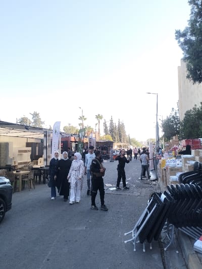 This image captures a daytime street scene, likely a market or exhibition, situated in the vicinity of the Homs National Museum in Homs, Syria. The sky is clear and bright.
On the left side of the asphalt pavement, the front section of a dark-colored SUV is visible. Adjacent to it, an outdoor seating area is set up with several square tables and wicker-style chairs, next to displays of furniture items under a canopy. Multiple white and green banners with Arabic script are positioned near these stalls.
In the mid-ground, three women walk together from left to right. The leftmost woman wears a black abaya and white hijab, carrying a dark bag. The middle woman also wears a black abaya and white hijab. The rightmost woman is attired in a white striped long shirt over white trousers with a white hijab. To their right, a young man in a light blue long-sleeved shirt and jeans stands looking towards the right. Nearby, a man in dark tactical attire, including a vest and dark trousers, stands, holding a device in his right hand. To his right, another man in a black long-sleeved shirt and black trousers is actively gesturing with both hands, his mouth open as if speaking or shouting, directed towards the right.
On the right side of the street, numerous stacks of black plastic folding chairs are displayed. Behind them, multiple cardboard boxes, some open, reveal white and colorful household items such as dishes and bowls. A man in a gray shirt stands near these items. Further down the street, additional individuals are visible, indicating an active public space.
The background features various trees, including palm trees. A prominent, light-colored, multi-story building with distinct vertical architectural elements, consistent with the Homs National Museum, stands on the far right. Streetlights are visible along the thoroughfare.